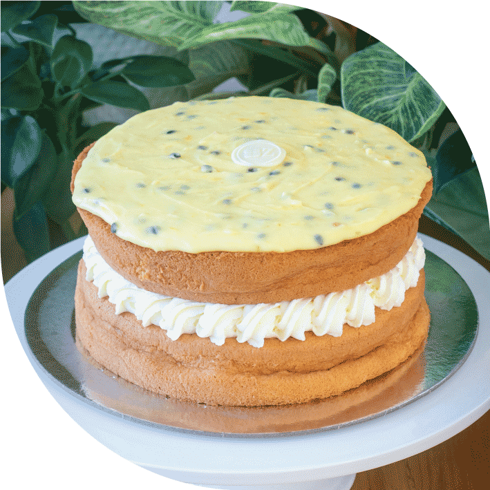 a golden sponge cake with generous cream filling topped with passionfruit icing, on a cake stand surrounded by plants