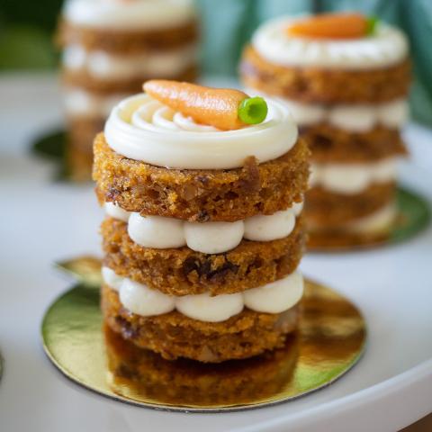 A close up of a single petit gateau carrot cake showing three layers with cream cheese icing and mini marzipan carrot decoration in front of two more carrot cakes in the background