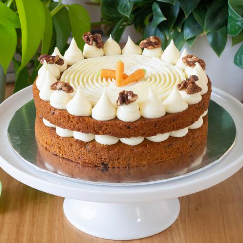 A two layered carrot cake decorated with cream cheese icing, caramelised walnuts and mini marzipan carrots on a cake stand in front of plants