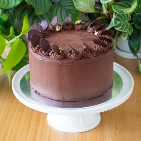 A tall chocolate covered prinzregententorte cake on a white cake stand in front of plants