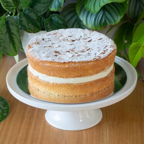 A golden sponge cake with generous cream filling topped with almonds and icing sugar, on a white cake stand in front of plants