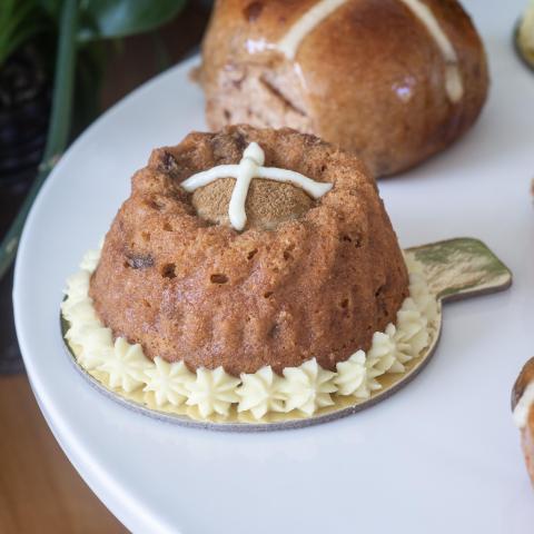 A close up of a Hot Cross Bundt with visible white cross on top and cream piping around the base with a Hot Cross Bun in the background