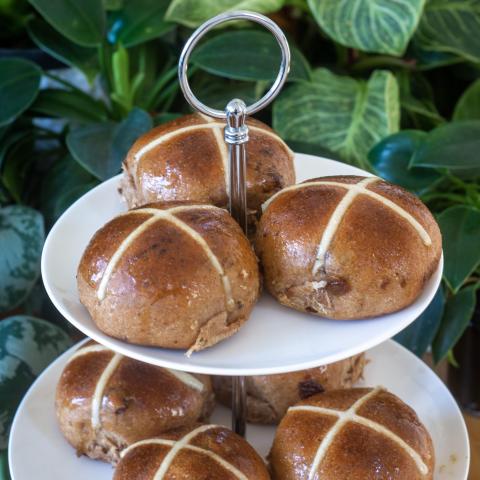 Several brown hot cross buns with white crosses on top sitting on a two-tiered white cake stand in front of greenery