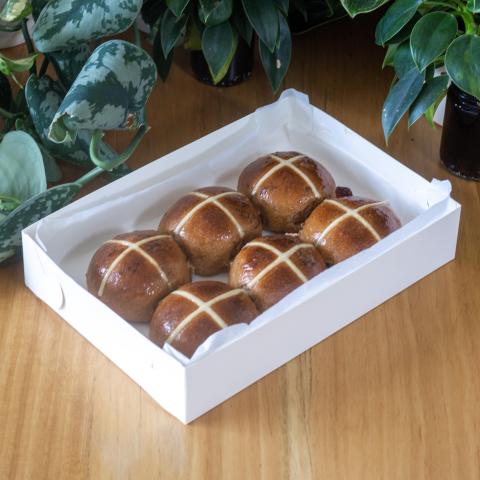 Six brown hot cross buns with white crosses on top in a white box in front of greenery