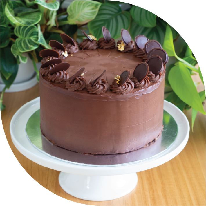 A tall chocolate covered prinzregententorte cake on a white cake stand in front of plants