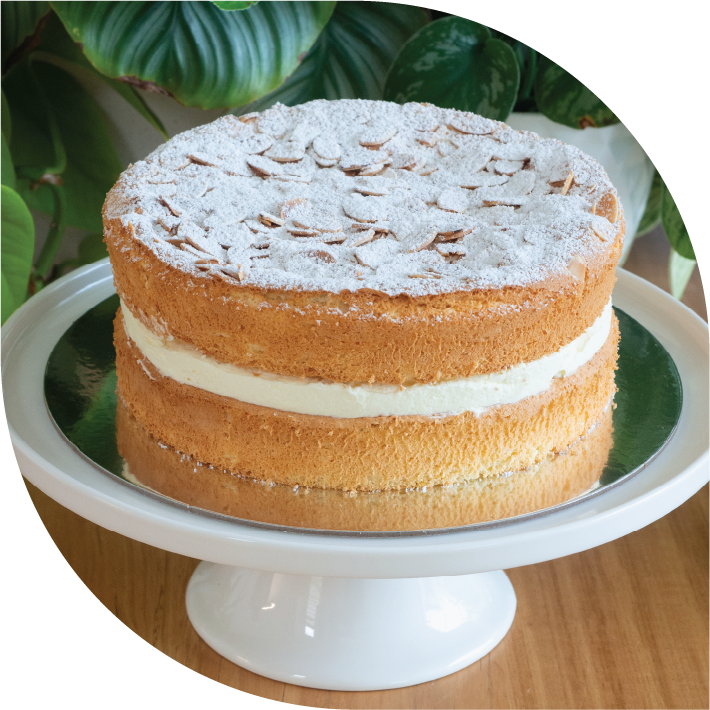 A golden sponge cake with generous cream filling topped with almonds and icing sugar, on a white cake stand
