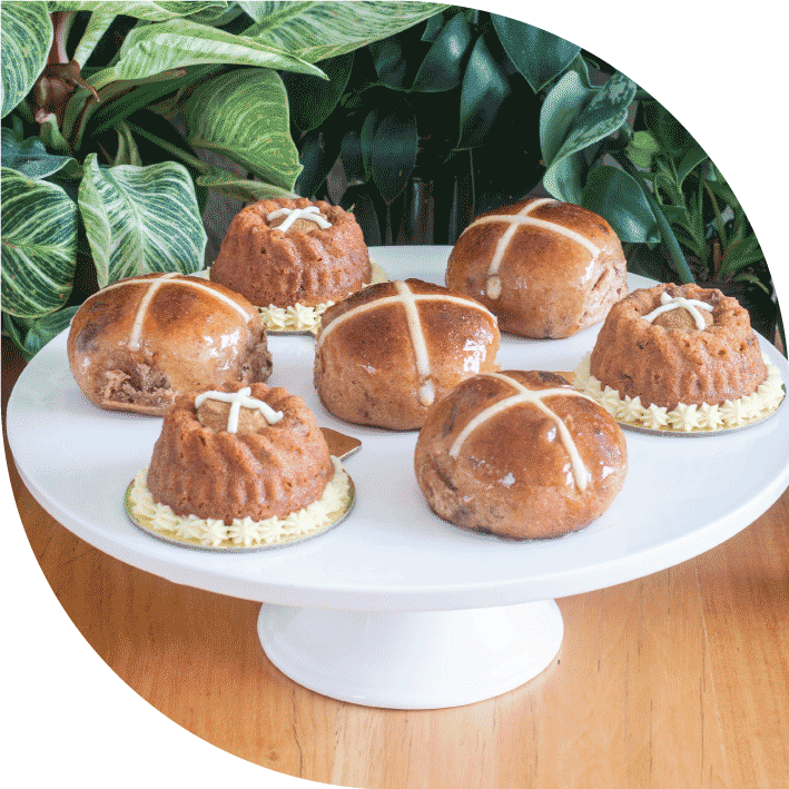 a white cake stand with four brown hot cross buns and three brown hot cross mini bundts all with white crosses on top, in front of greenery